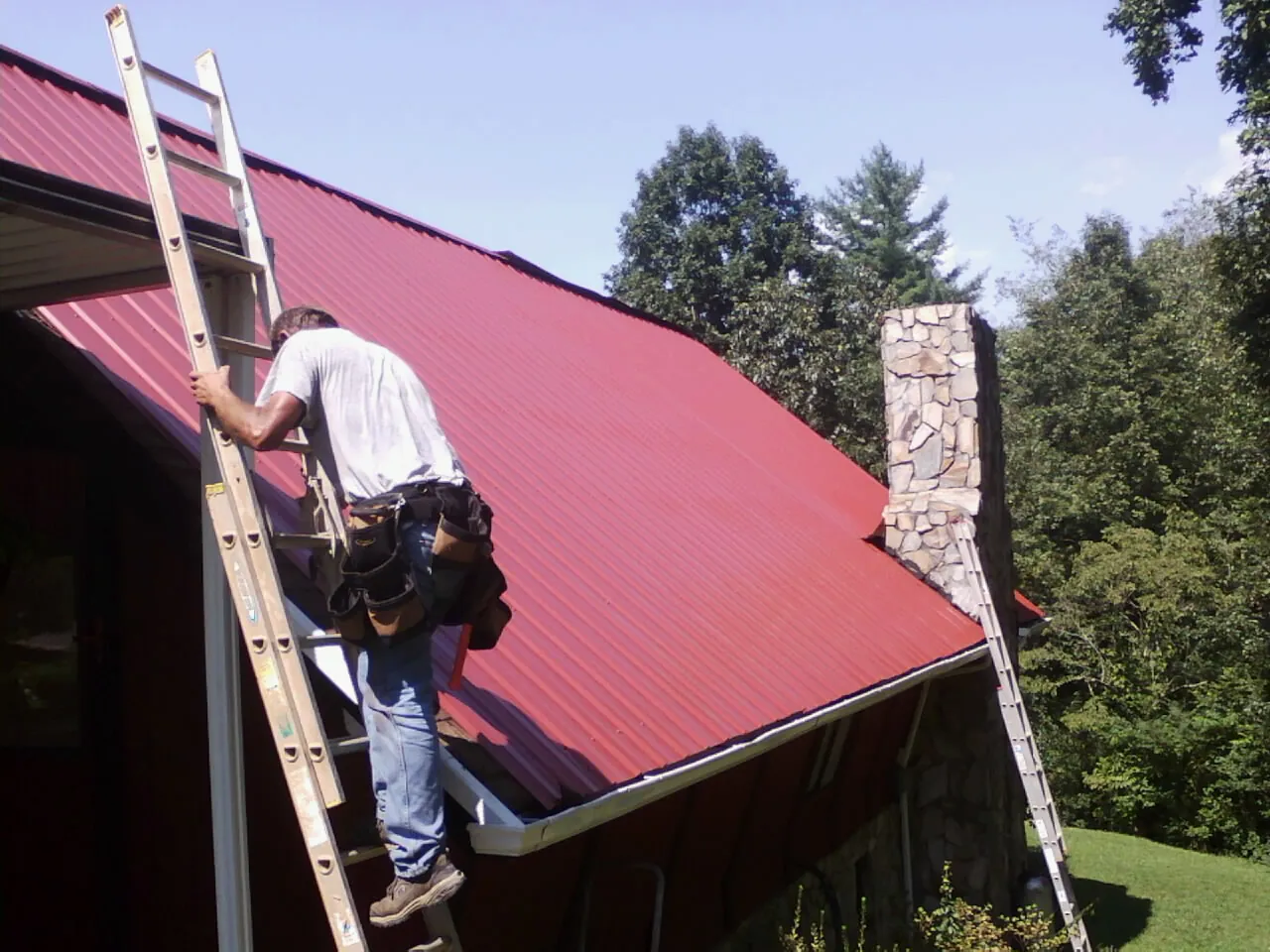Roofer on a ladder beside a red metal roof and stone chimney — Soffit & Fascia Repair by Roofing Brothers in Blairsville